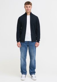 Donker marineblauwe ritsvest met ribbelstructuur; gedragen over een wit T-shirt. Gecombineerd met lichtblauwe jeans en witte sneakers met zwarte strepen.