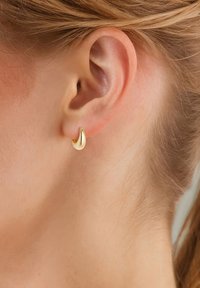 Zlato obarvani uhani v obliki obroča, ukrivljenega dizajna, sijoče površine, majhne velikosti, pritrjeni na ušesni mečici, z gladko teksturo in elegantno konturo.