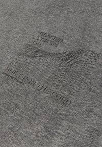 Grå bomullstyg med präglad text: "GLACIER SYSTEM OG" och "BUILT FOR THE COLD", med ett vågmönster för texturdetalj.