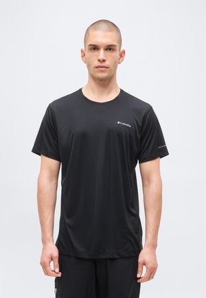 Mężczyzna z krótkimi włosami, ubrany w czarną koszulkę z krótkim rękawem marki Columbia i czarne spodnie, stojący na jednolitym tle.