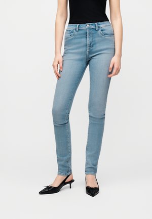 Frau trägt hellblaue Skinny-Jeans und schwarze Slingback-Pumps, steht vor einem schlichten weißen Hintergrund.