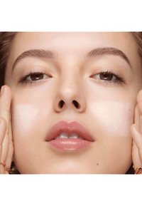 Nærbilde av et ansikt med glatt, lys hud. En tykk hvit krem er påført på kinnet. Naturlige lepper, lett rødme og velpleide øyenbryn.