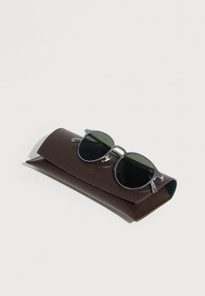 Στρογγυλά σκούρα γυαλιά ηλίου με μεταλλικούς βραχίονες που ακουμπούν σε κλειστή καφέ δερμάτινη θήκη γυαλιών πάνω σε λευκή επιφάνεια.