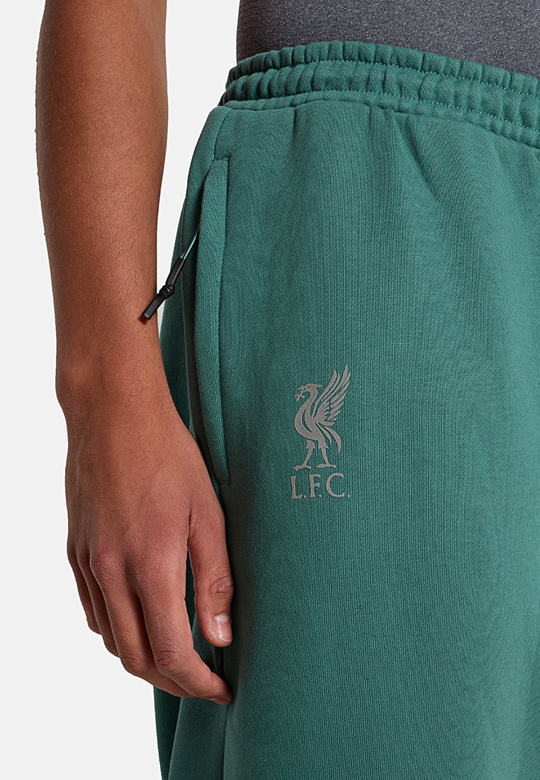 La mano sinistra di una persona poggia accanto a dei pantaloni da tuta verdi del Liverpool FC con cintura elasticizzata e tasca frontale, che mostrano il logo del liver bird del LFC.