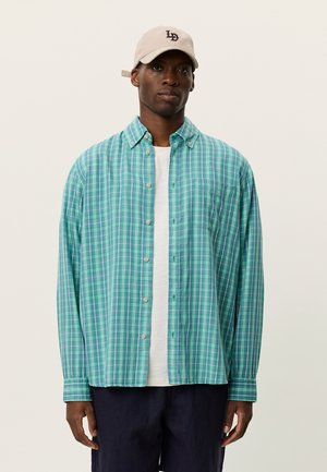 Koszula w zielono-niebieską kratę na guziki o luźnym kroju, z lekkiego materiału i długimi rękawami. Noszona na białym T-shircie, w połączeniu z ciemnymi szortami.