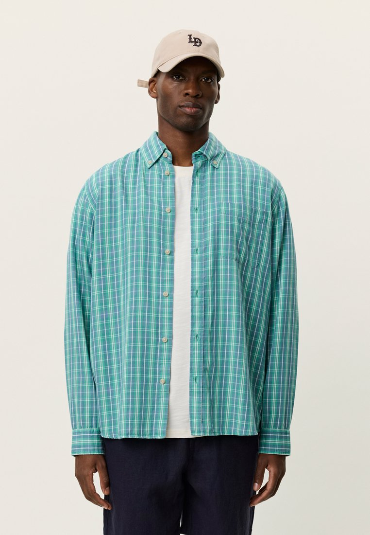Chemise à boutons à carreaux verts et bleus avec une coupe décontractée, un tissu léger et des manches longues. Portée par-dessus un t-shirt blanc, associée à un short foncé.