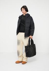 Νέος άνθρωπος που φοράει μια μαύρη μπουφάν, μαύρη μπλούζα, κρεμ φούτερ, καφέ μποτάκια, κρατώντας μια μαύρη τσάντα, στέκεται μπροστά σε έναν λευκό τοίχο.