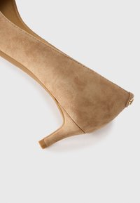 Sapato de salto alto em camurça bege com bico fino, salto esbelto e um pequeno elemento decorativo em tom dourado na parte de trás.