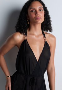 Robe noire en tissu transparent, dotée d'un décolleté en V profond, de bretelles ajustables avec des éléments en or, et d'une taille cintrée avec un lien.