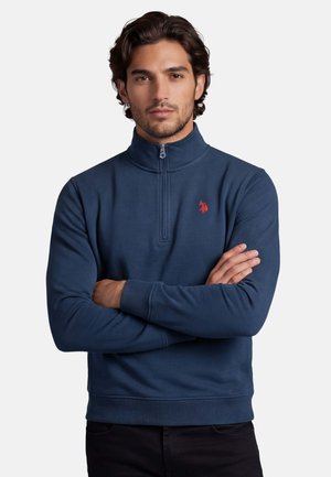 Mann trägt marineblaue Sweatshirt mit Viertel-Reißverschluss und rotem besticktem Logo, steht mit verschränkten Armen vor weißem Hintergrund.