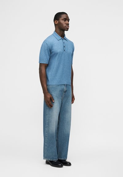 Άνδρας που στέκεται, φοράει μπλε κοντομάνικο πόλο, φαρδιά ξεθωριασμένα μπλε τζιν και μαύρα παπούτσια, μπροστά σε απλό λευκό φόντο.