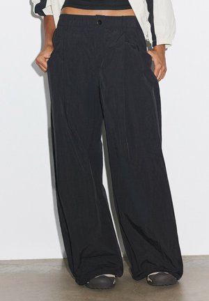 Persona che indossa pantaloni neri larghi, scarpe nere e un top nero corto con una giacca bianca, in piedi con le mani in tasca.