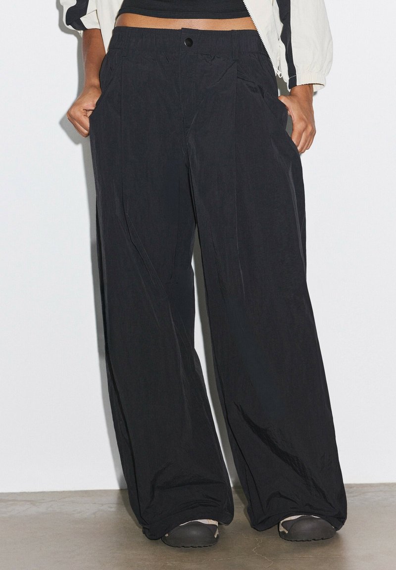 Personne portant un pantalon noir ample, des chaussures noires et un top noir court avec une veste blanche, debout les mains dans les poches.