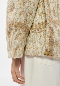 Tuvplāns ar personas roku blakus bēša un krēmkrāsas fakturētam tvīda žaketam ar pogu un baltām biksēm ar aizmugurējā kabatu.