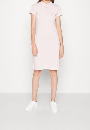 Frau trägt ein rosafarbenes Polo-Kleid in knieumspielender Länge und weiße Turnschuhe, steht vor einem schlichten weißen Hintergrund.