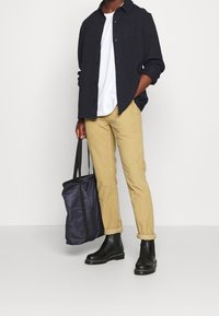 Camisa de botones azul marino sobre una camiseta blanca, combinada con pantalones beige claros, botas negras y un bolso tote gris oscuro.