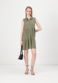Robe à manches courtes, verte olive, avec un devant boutonné et une jupe à volants. Présente un col et une coupe décontractée. Portée avec un petit sac noir.