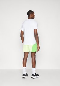 Λευκό αθλητικό μπλουζάκι με κοντά μανίκια, συνδυασμένο με ανοιχτό πράσινο σορτσάκι. Ορατό πόδι και λεπτομερείς ραφές στη μπλούζα και το σορτσάκι.