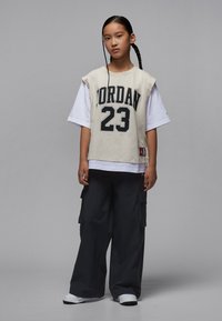 Top beige senza maniche con "Jordan 23" in nero, sovrapposto a una maglia bianca a manica lunga; abbinato a pantaloni cargo larghi scuri e sneakers bianche.