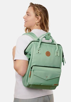 Femme aux cheveux ondulés portant une chemise blanche et un jean gris, avec un grand sac à dos vert menthe muni de poches avant et d'un écusson en cuir avec logo.