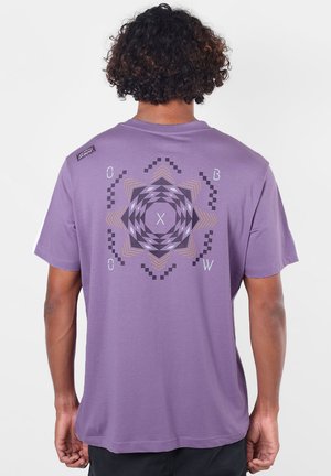 Personne aux cheveux bouclés portant un t-shirt violet avec un motif étoile géométrique et les lettres O, B, W et C au dos.