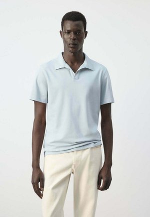 Joven de pie, vistiendo una camiseta tipo polo de manga corta azul claro y pantalones color crema, sobre un fondo blanco liso.