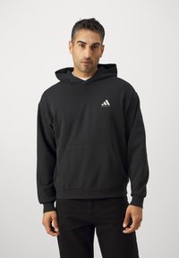 Hanorac negru cu glugă, realizat dintr-un amestec de bumbac, având un buzunar frontal și un logo alb Adidas pe pieptul stâng. Textură moale, croială lejeră.