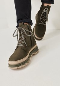 Grønne ruskindsankelstøvler med en kraftig beige sål, der har stribede snørebånd og en sidelukning for nem påtagning.