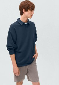 Námornícky modrá polo košeľa s krátkymi rukávmi, rebrovanými manžetami a klasickým golierom, v kombinácii s svetlo sivými denimovými šortkami. Hladká textúra látky.