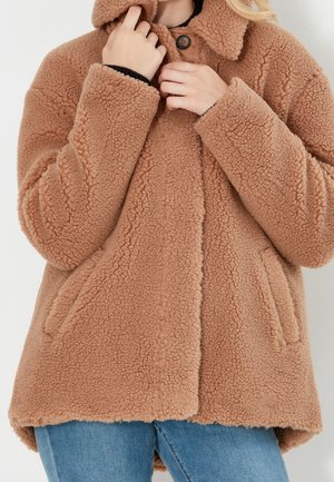 Person iført en tanfarvet, fluffy teddyfrakke, der knapper kraven, kombineret med blå jeans mod en ensfarvet lys baggrund.