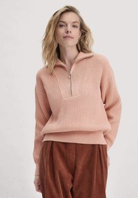 Helle rosa gestrickter Pullover mit einem Viertelreißverschluss-Kragen, strukturierten vertikalen Streifen und gerippten Säumen, kombiniert mit braunen Cordhosen.