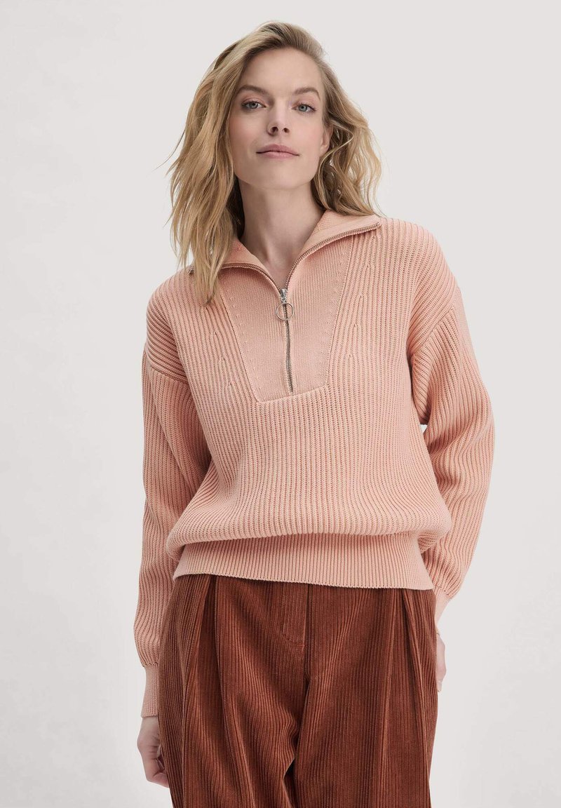 Helle rosa gestrickter Pullover mit einem Viertelreißverschluss-Kragen, strukturierten vertikalen Streifen und gerippten Säumen, kombiniert mit braunen Cordhosen.