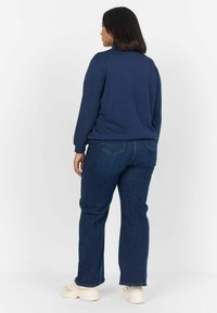 Sudadera azul marino con un cuello alto, mangas largas; combinada con jeans rectos de color azul oscuro y zapatillas crema, mostrando un look simple y casual.
