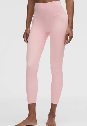 Kvinde iført lyserøde, tætsiddende leggings med høj talje, stående barfodet mod en ensfarvet hvid baggrund.