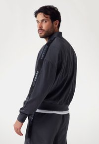 Black nylon bomber jacket with ribbed cuffs and collar, featuring a "GUESS" logo on the side. Smooth texture, casual fit.