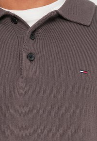 Nahaufnahme eines grauen Strick-Poloshirts mit drei schwarzen Knöpfen und kleinem roten, weißen und blauen Logo auf der Brust über einem weißen Unterhemd.