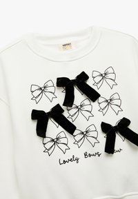 Biała bluza z czarnym nadrukiem w wzór kokardek oraz czterema przyszytymi czarnymi aksamitnymi kokardami. Pod spodem znajduje się napis "Lovely Bows Crew."