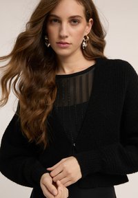 Musta lyhyt neuletakki teksturoidusta neuleesta, jossa on syvä V-pääntie ja leveät hihat, kerroksittain läpikuultavan mustan yläosan päällä, jossa pystysuoria helmiäiskoristeita.