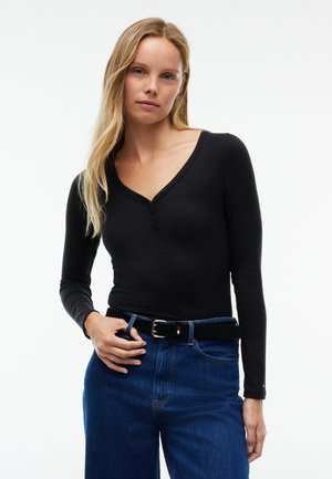 Femme aux longs cheveux blonds portant un haut noir moulant à manches longues et un jean bleu avec une ceinture noire, debout devant un fond uni.