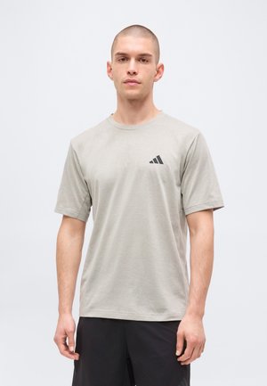 Jeune homme aux cheveux courts portant un t-shirt Adidas gris clair et un short noir, debout devant un fond blanc uni.