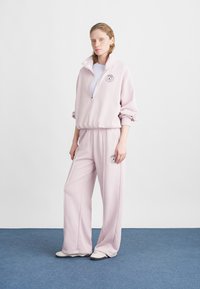 Lyserød, lynlås sweatshirt med elastiske manchetter, parret med matchende vide sweatpants, som har et logo på brystet og på bukserne. Glat tekstur.