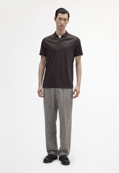 Brązowa koszulka polo wykonana z gładkiego materiału, w zestawie z jasnoszarymi spodniami o teksturowanej fakturze. Model stoi przed gładkim białym tłem.