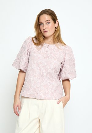 Žena svjetlosmeđe kose nosi teksturiranu blijedo ružičastu bluzu s naboranim rukavima i kremaste široke hlače, stoji s jednom rukom u džepu.