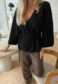 Top negru stil wrap, cu decolteu în V și mâneci largi, purtat cu pantaloni maronii conici. Textură moale, talie strânsă cu un detaliu de sfoară.