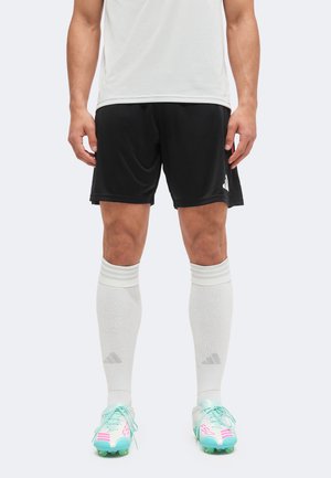 Männlicher Athlet trägt schwarze Shorts, ein weißes Shirt, weiße knielange Socken und hellblaue Fußballschuhe mit pinken Akzenten und steht aufrecht.