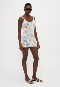 Vestido de ganchillo blanco con patrones de estrellas de mar y espirales de colores, adornado con acentos multicolores. Combinado con gafas de sol negras y sandalias.