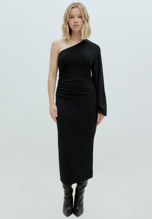Robe noire ajustée avec un design asymétrique ; une manche et des détails froncés à la taille. Portée avec des bottes noires jusqu'aux genoux.