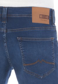 Granatowe dżinsy z klasycznym krojem. Wśród cech znajdują się brązowa skórzana metka oraz pomarańczowe przeszycia wzdłuż kieszeni.