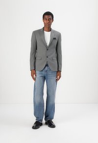 Grå skreddersydd blazer med spalteklaffer, en enkelt brystlomme, sammen med blå vide bukser og svarte sko. En enkel hvit T-skjorte under.