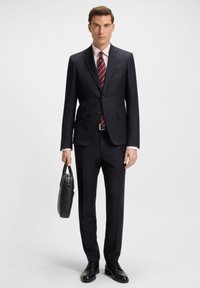 Homme en costume sombre à fines rayures, cravate rouge à rayures, et chemise rose tenant une mallette en cuir noir, debout devant un fond blanc.